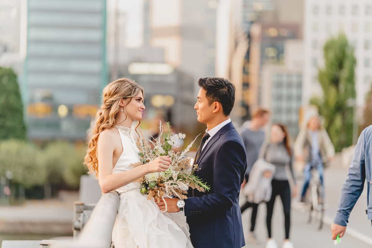 Braut mit Brautstrauß in A-Linie Brautkleid. Bräutigam mit schwarzem Anzug. Urbane Hochzeit in Frankfurt am Main zwischen den Hochhäusern.